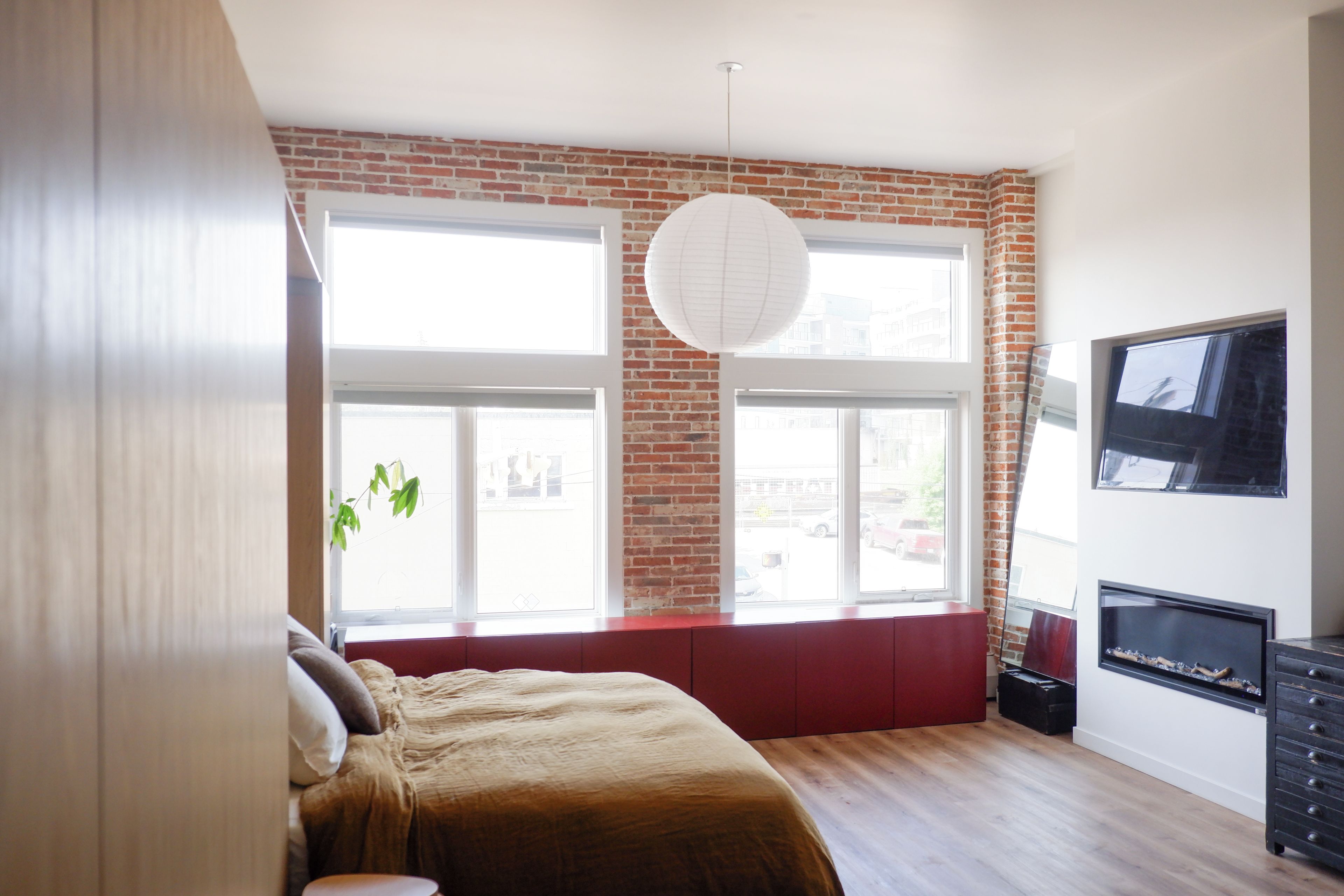 A bedroom with a brick wall, large windows, a bed, a TV, and a fireplace.