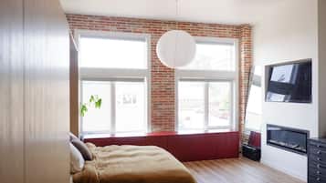 A bedroom with a brick wall, large windows, a bed, a TV, and a fireplace.