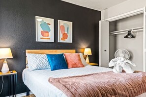 A bedroom with a bed, two bedside lamps, a bedside table, a wall-mounted fan, and abstract artwork. - Stylish & Comfy Downtown Gem ~ Workspace ~ Parking (Irving)