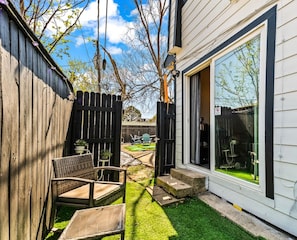 A residential backyard with a wooden fence, a paved patio, and a sliding glass door leading to a green lawn. - Stylish & Comfy Downtown Gem ~ Workspace ~ Parking (Irving)