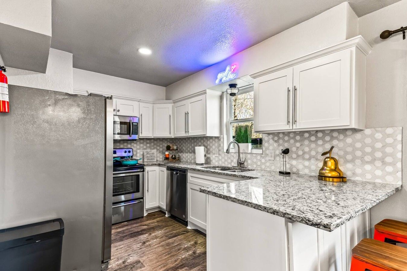 A modern kitchen with white cabinets, a granite countertop, stainless steel appliances, and a neon sign.