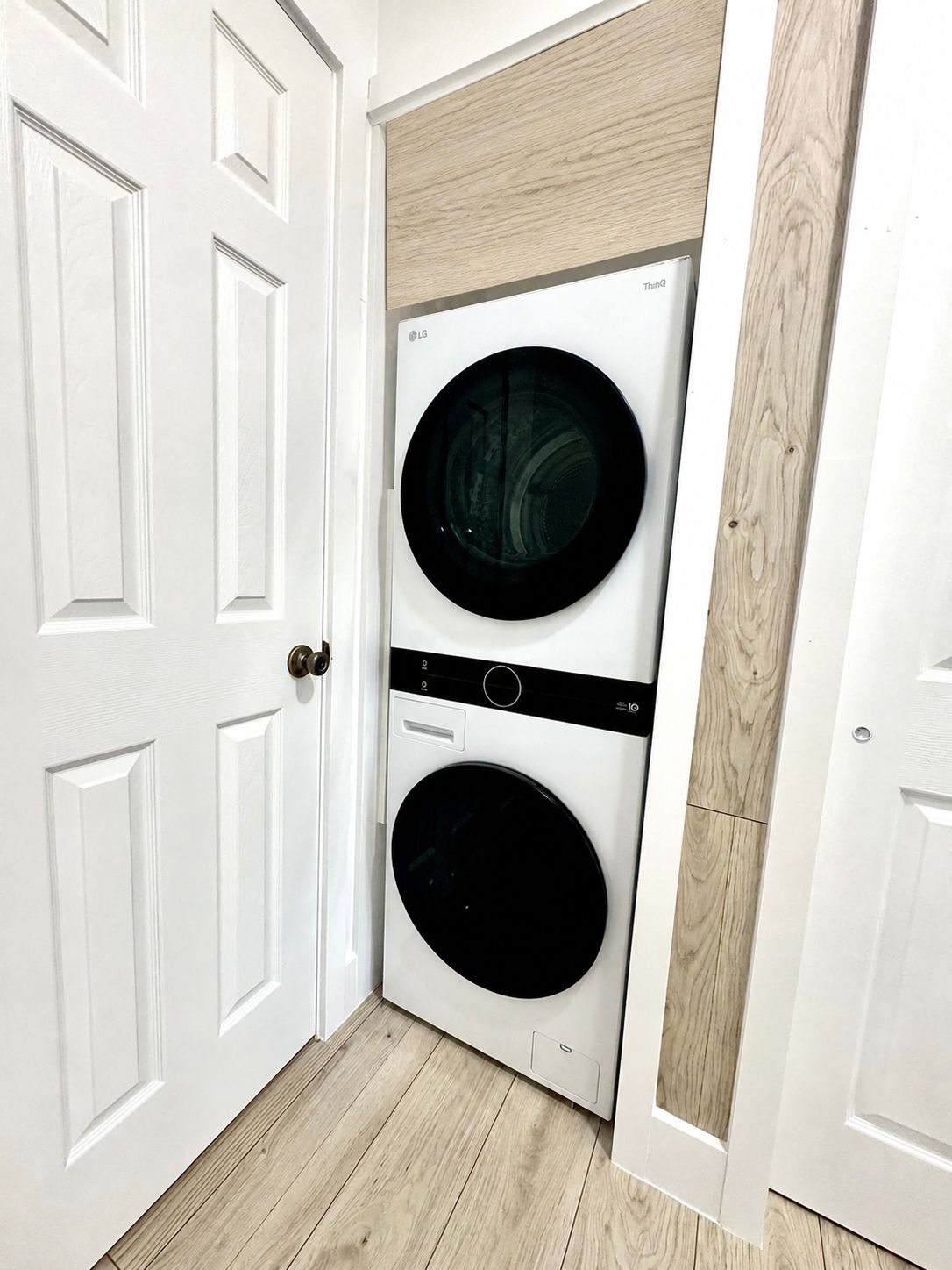 Una sala de lavandería con dos lavadoras apiladas, una puerta blanca y piso de madera.