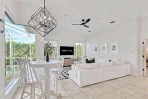 A modern living room with a white sofa, a dining table with chairs, a ceiling fan, and a TV screen displaying 'Home Team'. - Waterfront Oasis • Pool •  Private Dock • Firepit (Marco Island)