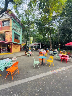 Un café en plein air avec des chaises et des tables aux couleurs vives, entouré d’arbres et d’immeubles.