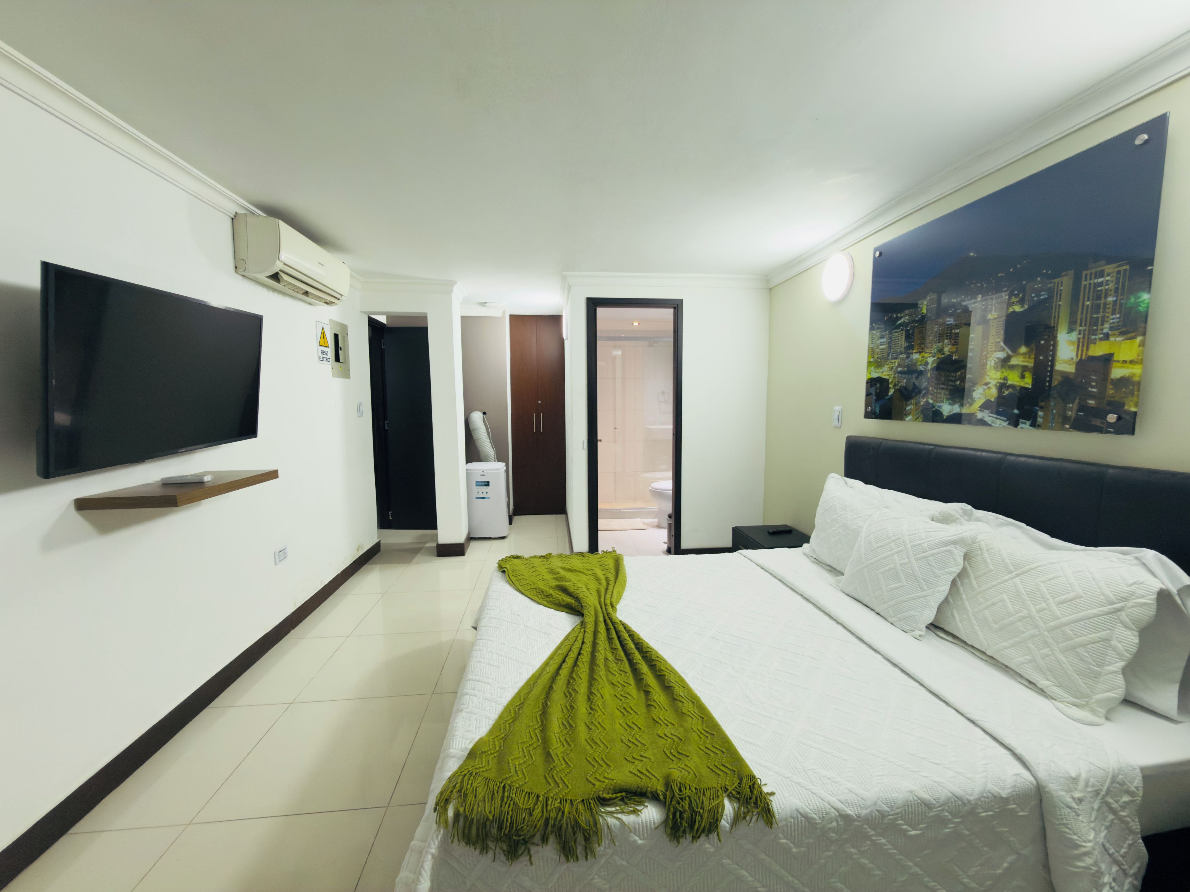 Ein Hotelzimmer mit einem Bett, einem Fernseher, einem Stadtbildgemälde und einem durch eine offene Tür einsehbaren Badezimmer.