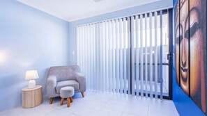 A room with a grey armchair, a small round table with a lamp, and a large window with vertical blinds. - Lakelands Escape Western Australia (Lakelands)