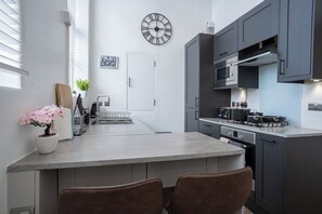 A modern kitchen with a central island, dark cabinets, and a wall-mounted clock. - 2BR Flat - Greenwich - Parking - Elizabeth Line (Greater London)
