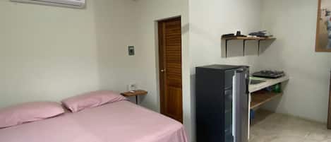 A bedroom with a bed, a ceiling fan, an air conditioner, a refrigerator, and a small kitchenette area with a shelf and a stove.