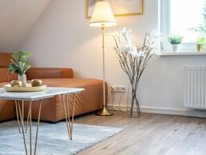 Een moderne slaapkamer met een bank, een nachtkastje met een lamp, een vaas met bloemen en een raam met planten.