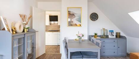 A modern kitchen with white cabinets, a dining area with a grey table and chairs, and a clock on the wall.