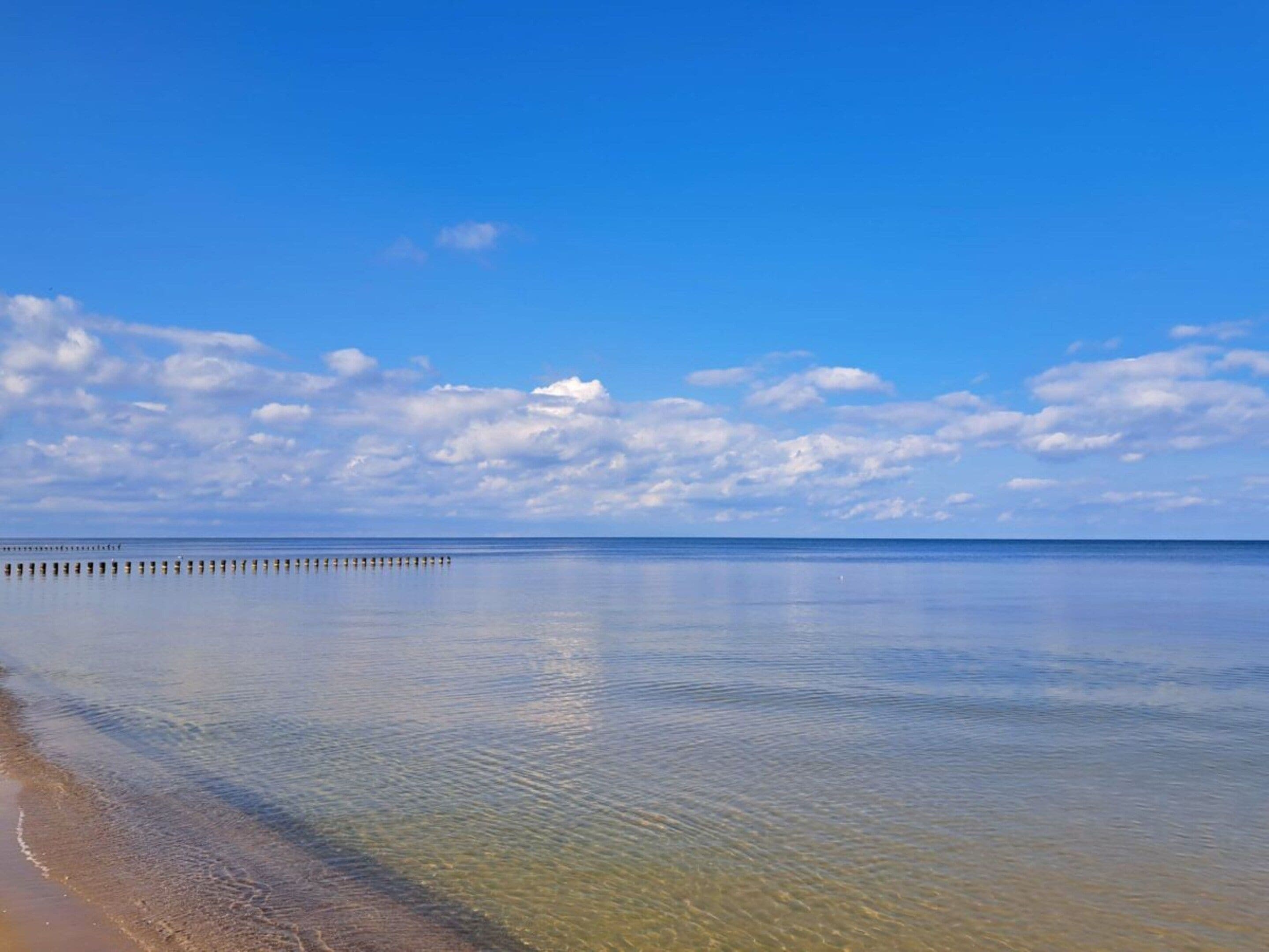 A calm beach with clear water, a wooden pier extending into the sea, and a partly cloudy sky.