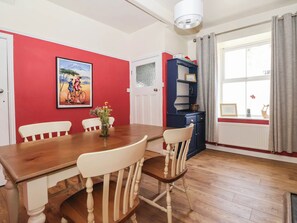 A dining area with a wooden table and chairs, a red wall with a framed picture, a blue cabinet with decorative items, and a window with curtains. - Bowdery (Harrogate)