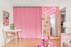 A room with a pink curtain, a round table with a chair, a potted plant, a mirror with floral wallpaper, and a pink sofa. - Glam Society Music Row-IG Worthy-Outdoor Lounge (Nashville)