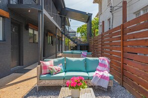 A modern outdoor seating area with a sofa and cushions, a small table with a flower vase, and a wooden fence. - Glam Society Music Row-IG Worthy-Outdoor Lounge (Nashville)