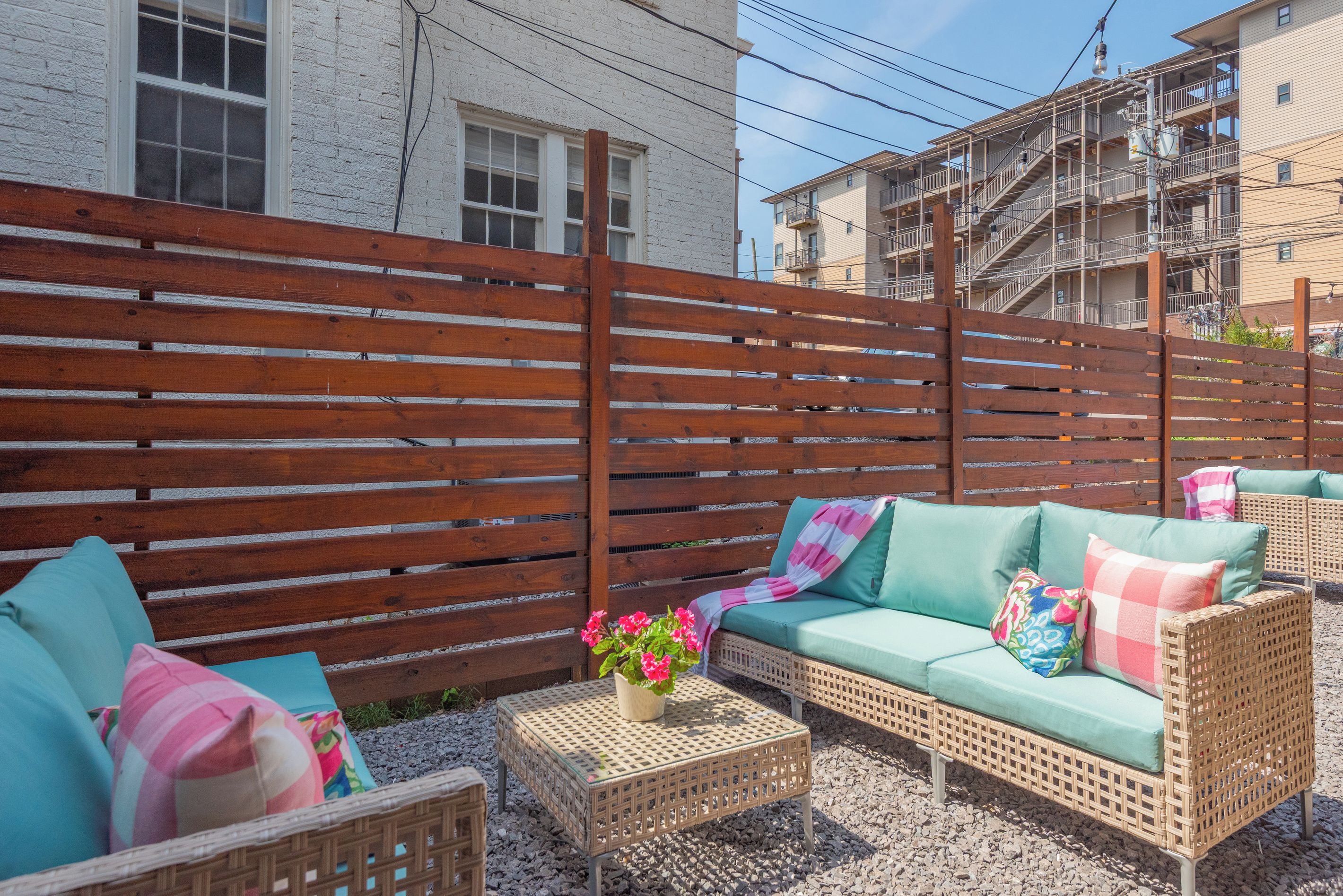 Ein Terrassenbereich mit Holzlattenzaun, Korbmöbeln und einem kleinen Tisch mit Blumenarrangement.