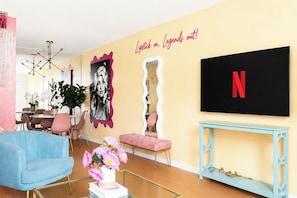 Een woonkamer met een televisie, een roze muur met een citaat, een blauwe bank en een roze poef.