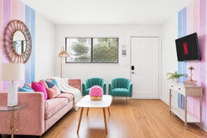 Ein Wohnzimmer mit einem rosa Sofa, Salbeigrün-Armstühlen, einem Glastisch und einem an der Wand montierten Fernseher.