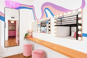 Ein Zimmer mit Schminkspiegel, Beleuchtung, einem Etagenbett und rosafarbenen Hocker.