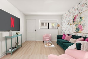 Ein Wohnzimmer mit einem großen Flachbildfernseher, einem rosa Sessel, einem grünen Sofa und floraler Tapete.
