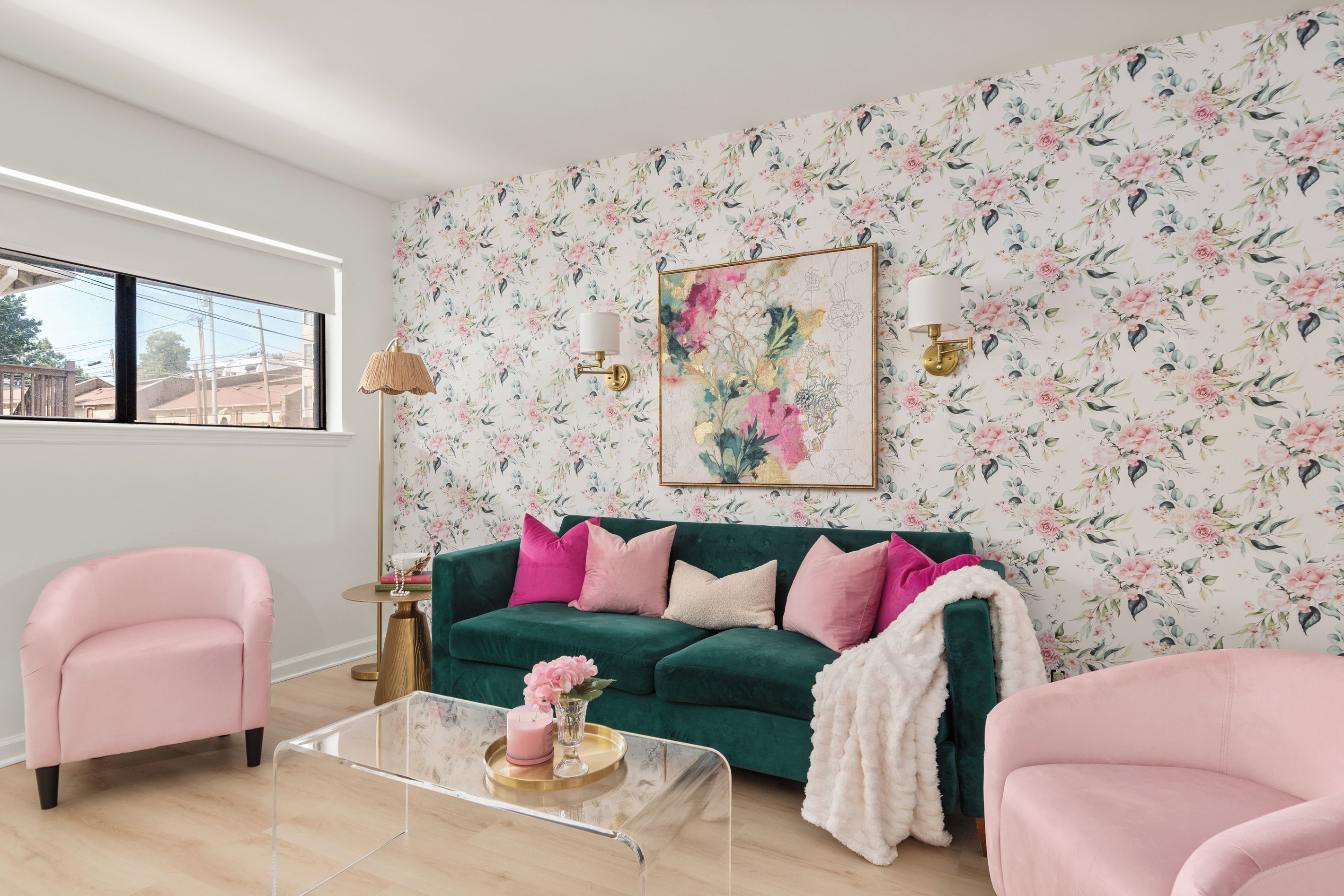 A living room with a floral wallpaper, a green sofa with pink cushions, a pink armchair, and a clear coffee table with a candle and flowers.