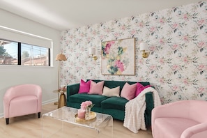 Un salon avec un papier peint à motifs floraux, un canapé vert agrémenté de coussins roses, un fauteuil rose et une table basse en verre sur laquelle sont posés une bougie et des fleurs.