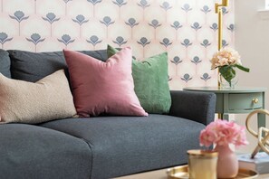 Ein graues Sofa mit mehreren Kissen in verschiedenen Farben, ein kleiner Beistelltisch mit einem Blumenstrauß und ein gemustertes Hintergrundpapier.