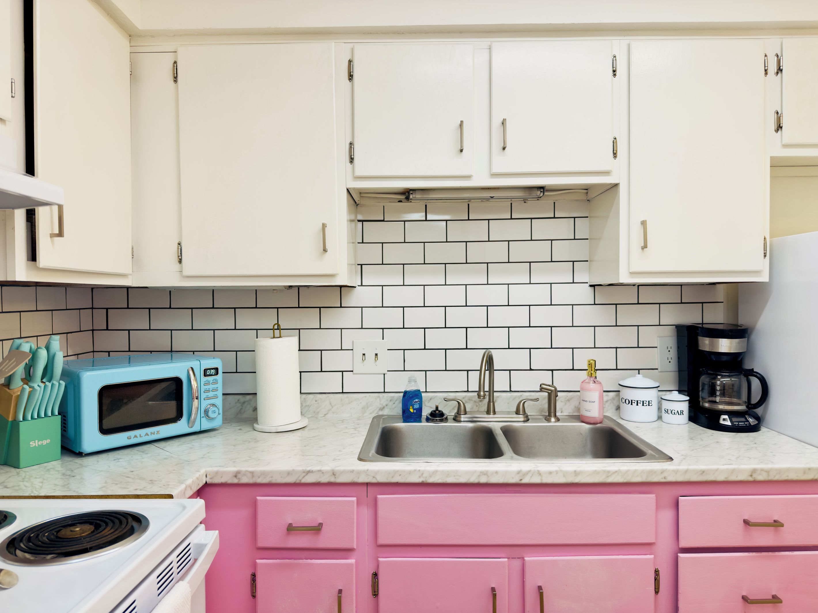 Una cocina con armarios blancos, una encimera rosa, un microondas azul, un lavabo y una cafetera.