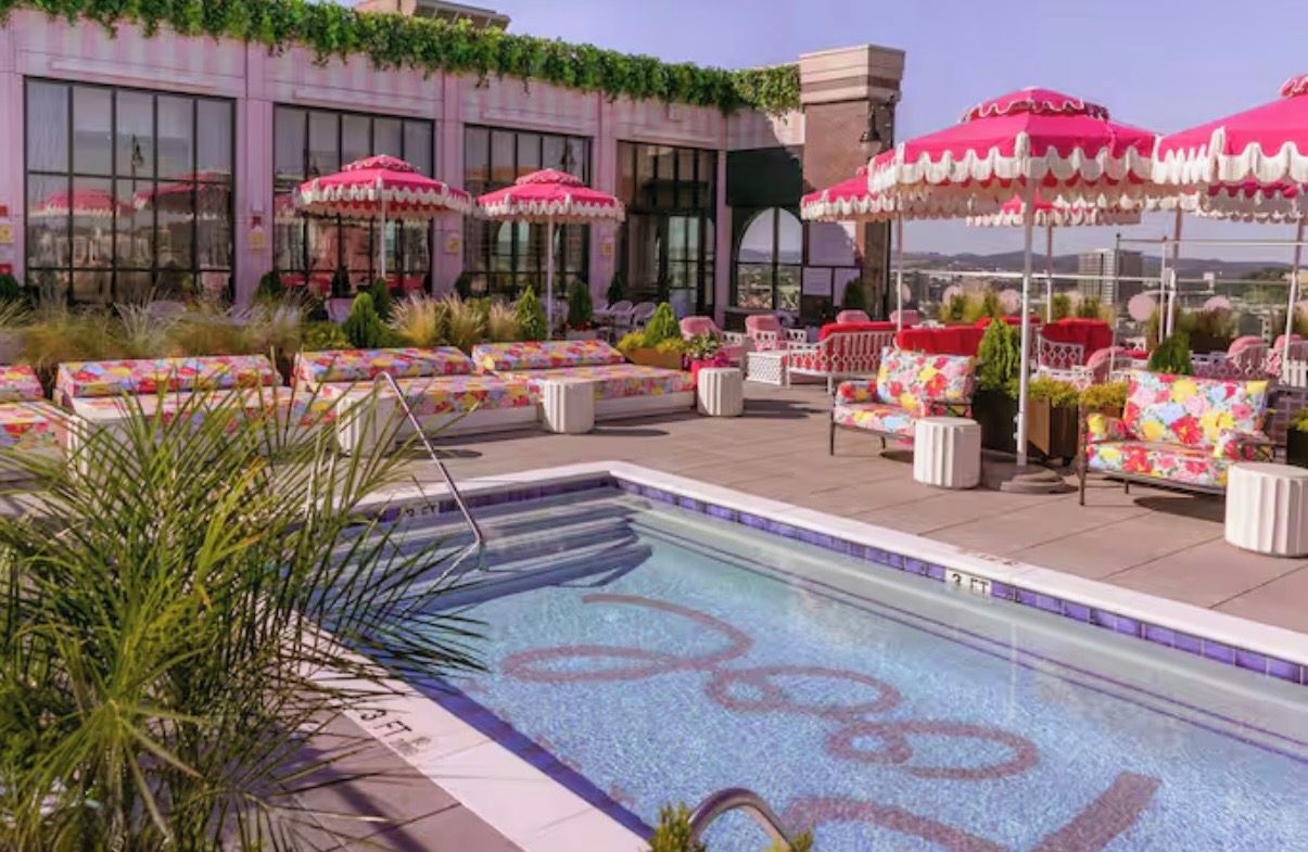 A rooftop pool area with lounge chairs and umbrellas, surrounded by a building with large windows.