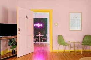 Een kamer met een roze muur, een tekst in een lijst aan de muur, een deur met een neonbord, een televisie en een plant.