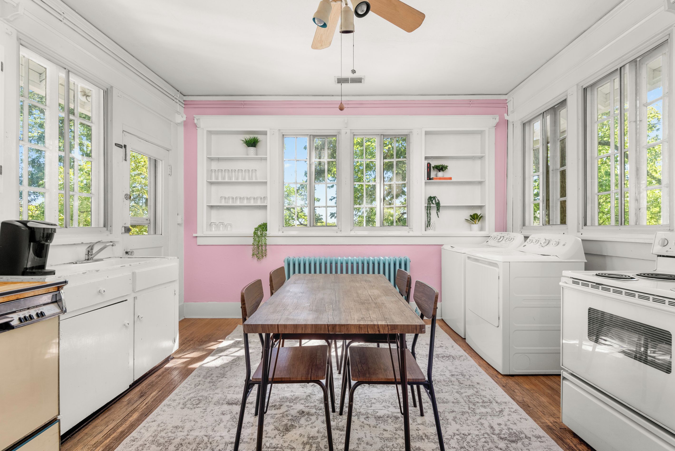 Una cocina con una mesa de madera, sillas, armarios blancos, una pared rosa y una ventana con vistas a árboles.
