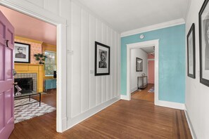 Un couloir aux murs recouverts de panneaux blancs, doté d’un plancher en bois et d’une porte donnant sur une pièce dont un mur est peint en rose et qui contient une cheminée.
