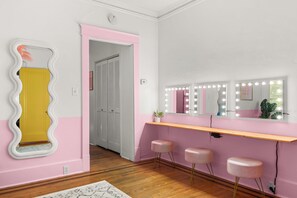 Une pièce avec une coiffeuse, un miroir avec éclairage, une porte et un mur rose.
