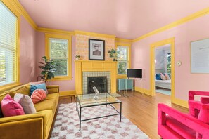 Ein Wohnzimmer mit gelber Couch, rosa Sessel, Glastisch, Kamin und Fernseher.