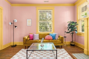 A room with a yellow sofa, a glass coffee table, a pink wall, and a window with blinds. - Taylor's Music Row Glam Near Broadway IG Worthy (Nashville)