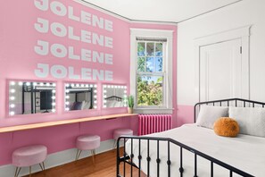 Ein Schlafzimmer mit rosafarbenen Wänden, einem Bett, einem Fenster mit Aussicht und einem Schminktisch mit Spiegeln und Beleuchtung.