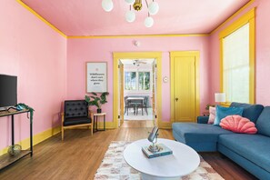 Ein Wohnzimmer mit rosaroter Wand, einem blauen Sofa, einem schwarzen Sessel, einem Couchtisch und einem Fernseher.