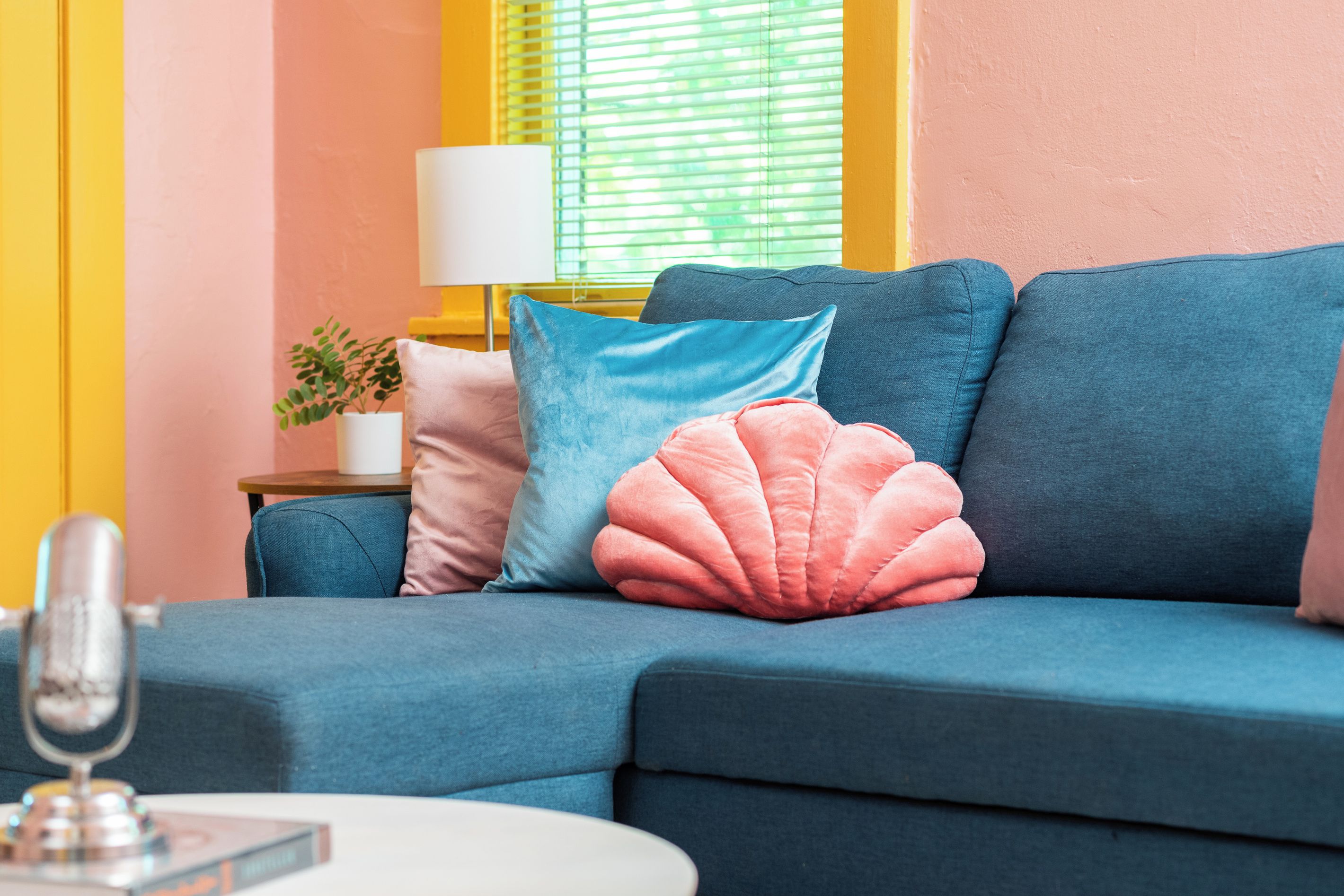 A blue sofa with pink and blue cushions, a white lamp, and a small plant on a side table.