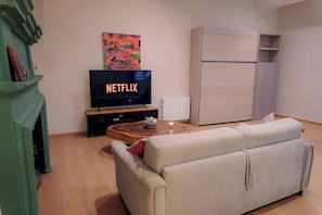 Ein Wohnzimmer mit einem Fernseher, der 'NETFLIX' anzeigt, einem Holztisch, einem beigefarbenen Sofa und einem weißen Schrank.