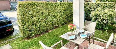 A patio with wicker furniture, a table set for dining, and a neatly trimmed hedge.