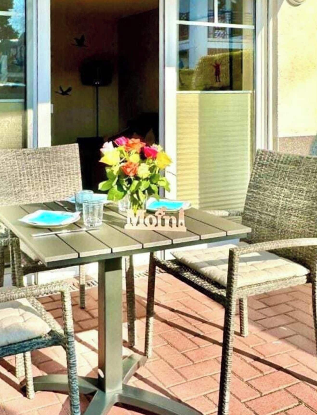 An outdoor patio with a table set for two, featuring wicker chairs and a vase with flowers.