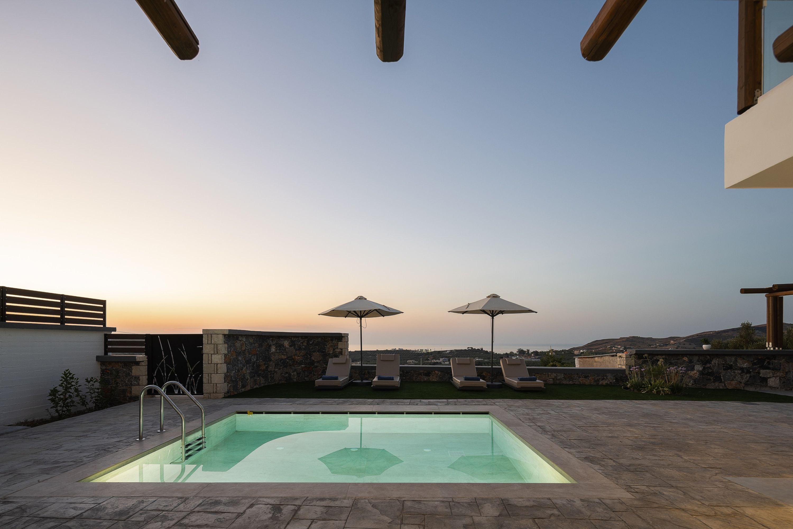 A swimming pool with lounge chairs and umbrellas, surrounded by stone walls and a view of the sea at sunset.