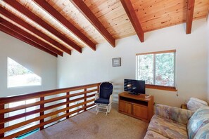 Ein Zimmer mit Holzboden, einem Fernseher auf einem Holzständer, einer Couch, einem Sessel und einem Fenster mit Blick auf Bäume.
