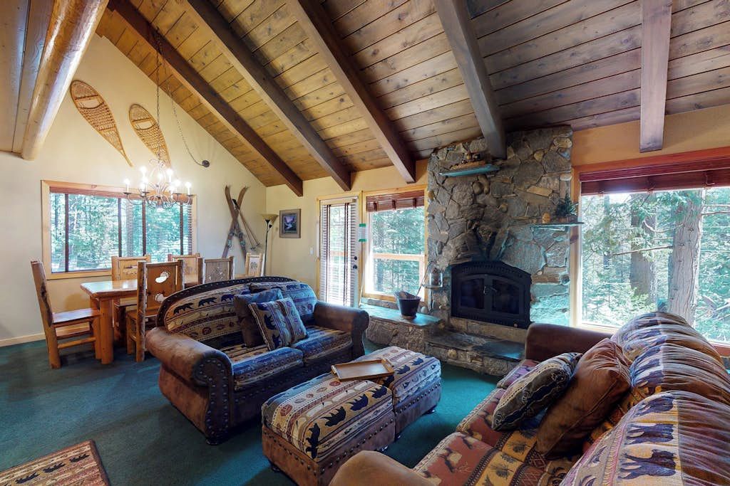 A cozy living room with a stone fireplace, wooden beams, and a large window overlooking trees.
