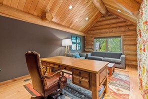 A log cabin interior with a wooden desk, leather chair, sofa, and a window with a view of trees. - Willow Cove Log Cabin (Bass Lake)