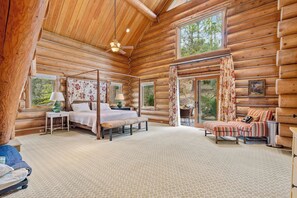 A log cabin bedroom with a four-poster bed, a sitting area with a striped sofa, and a large window offering a view of the outdoors. - Willow Cove Log Cabin (Bass Lake)