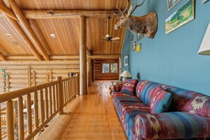 A log cabin interior with a wooden ceiling, a mounted deer head, a colorful sofa, and framed pictures on the walls. - Willow Cove Log Cabin (Bass Lake)
