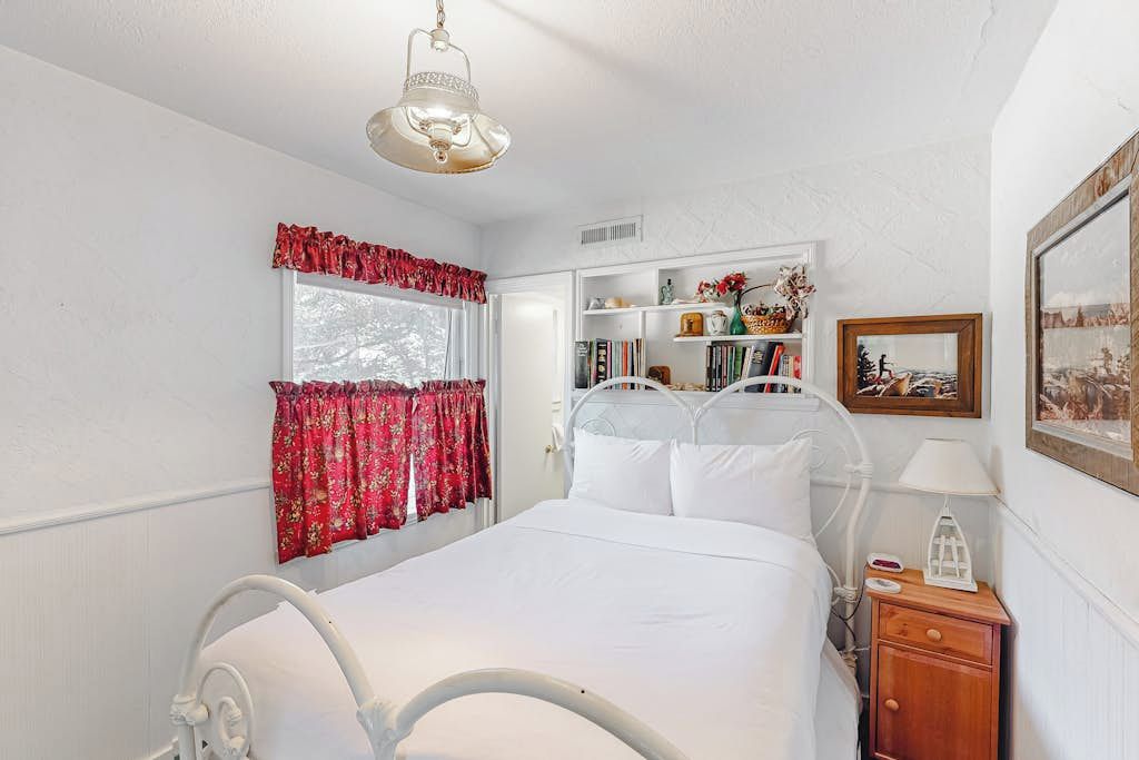Une chambre à coucher avec un lit, une table de chevet, une lampe, une fenêtre avec des rideaux, une bibliothèque et un tableau encadré au mur.