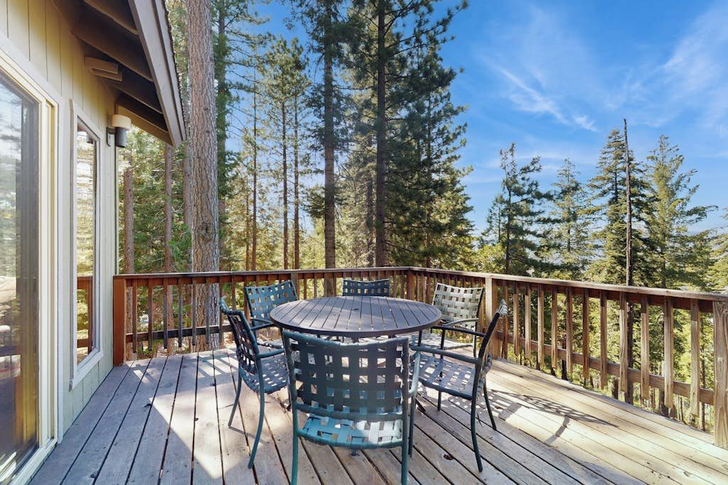A wooden deck with a round table and chairs, surrounded by tall trees.