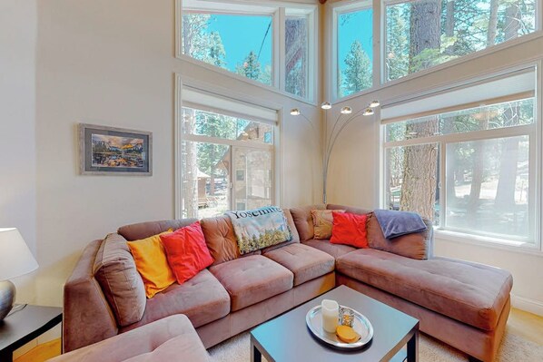 A living room with a brown sectional sofa, a coffee table, and large windows offering a view of trees.