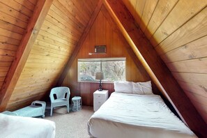 Un dormitorio acogedor en el ático, con techo inclinado, dos camas, una silla, una mesita de noche y un ventanal con vista a árboles.